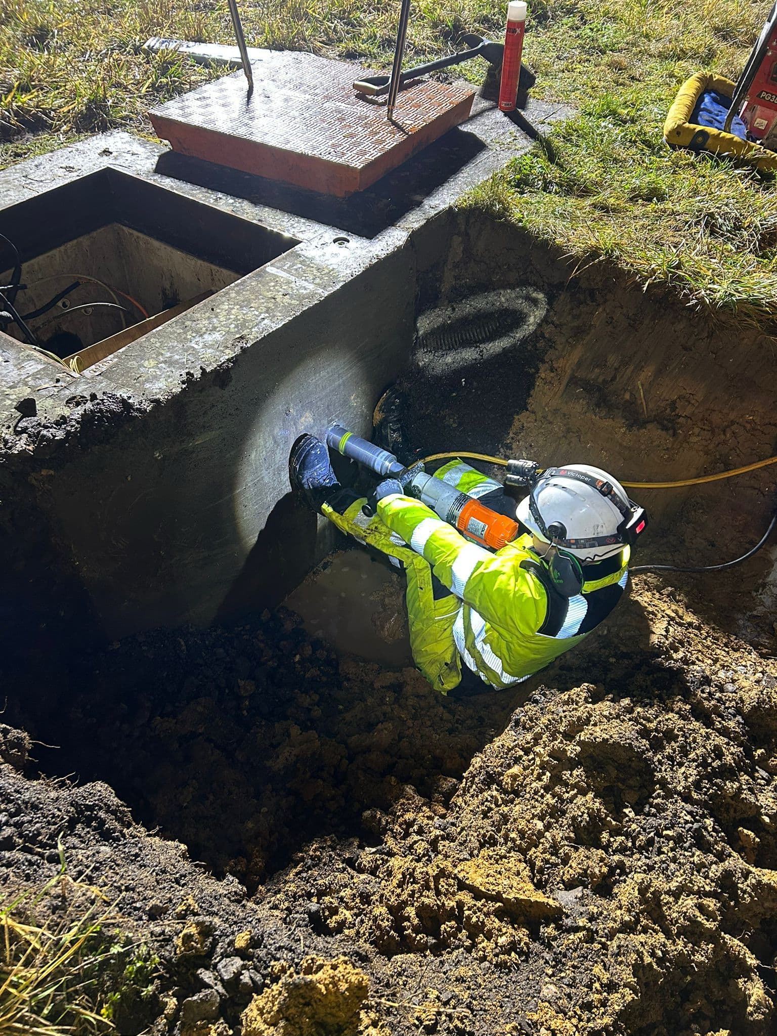 Worker in hi-vis and helmet kneeling in an excavated pit using a core drill on a concrete chamber in daylight