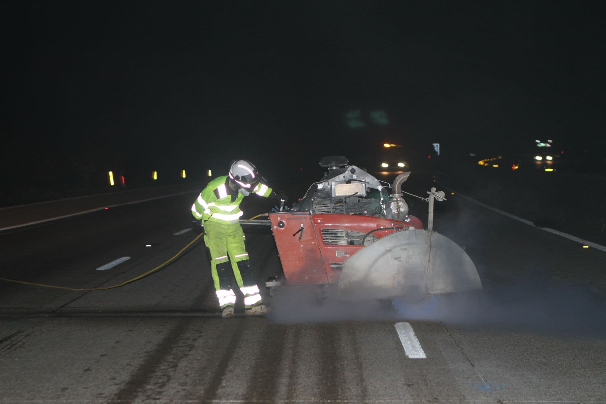 Worker in green hi-vis crouching over a red floor saw on a road at night with dust around the blade