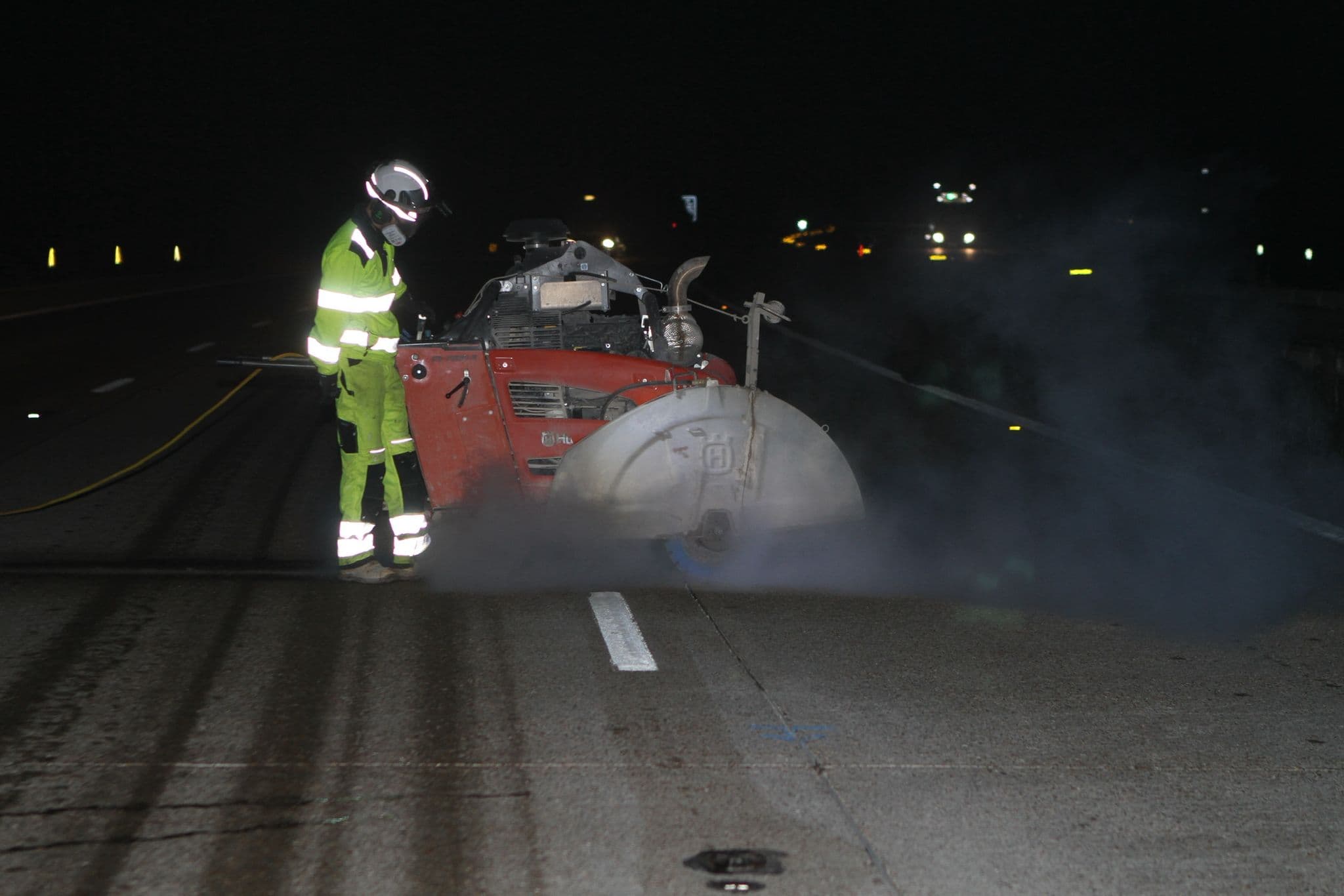 Worker in green hi-vis and helmet operating a red floor saw at night with dust around the cutting blade