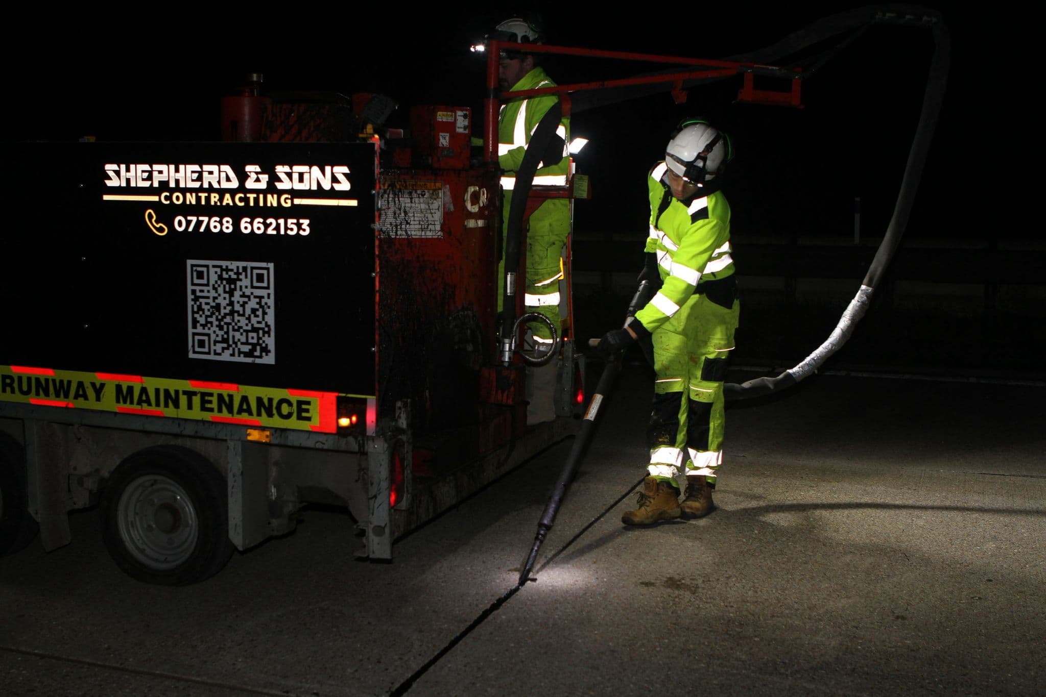 Worker in hi-vis and hard hat at night operating a lance beside a Shepherd and Sons Contracting runway maintenance trailer