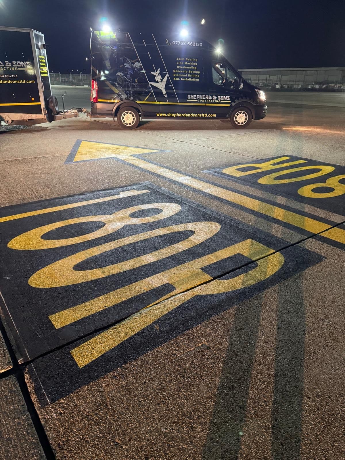 Freshly painted yellow STOP road marking and arrow at night beside a Shepherd and Sons Contracting branded van and trailer