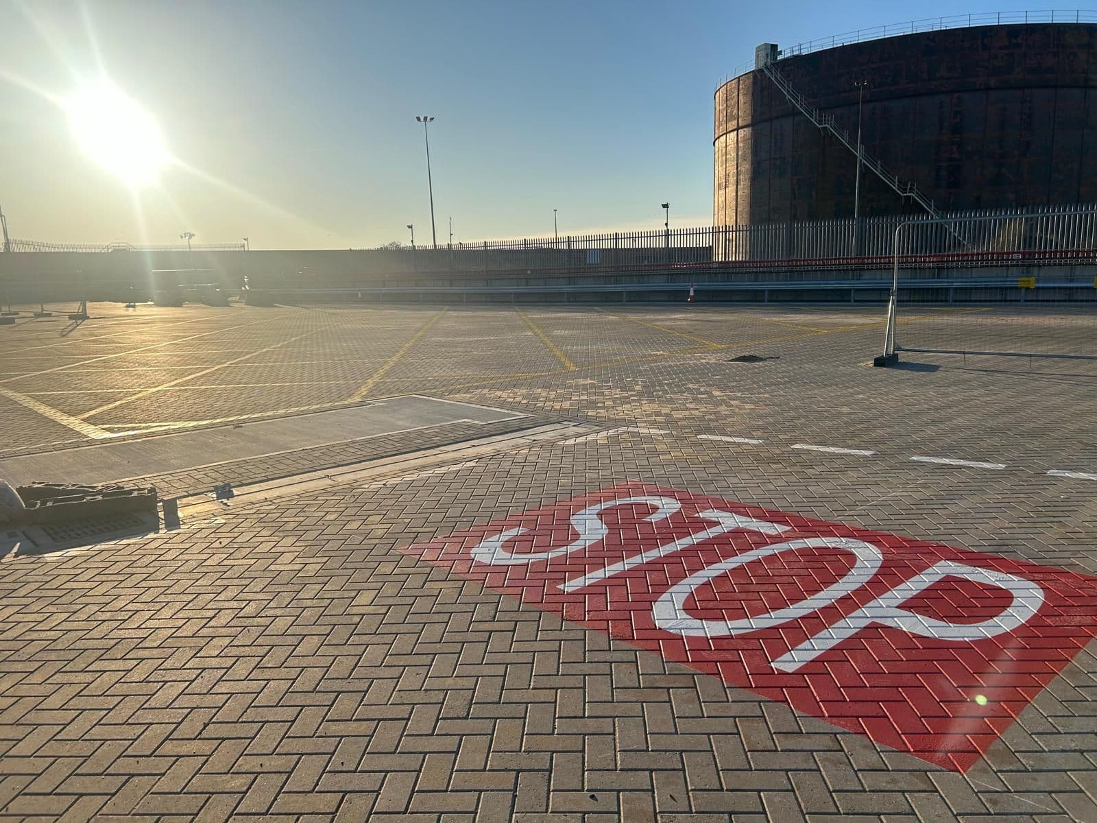 Large red STOP road marking painted on block-paved ground at a fenced yard in bright sunlight