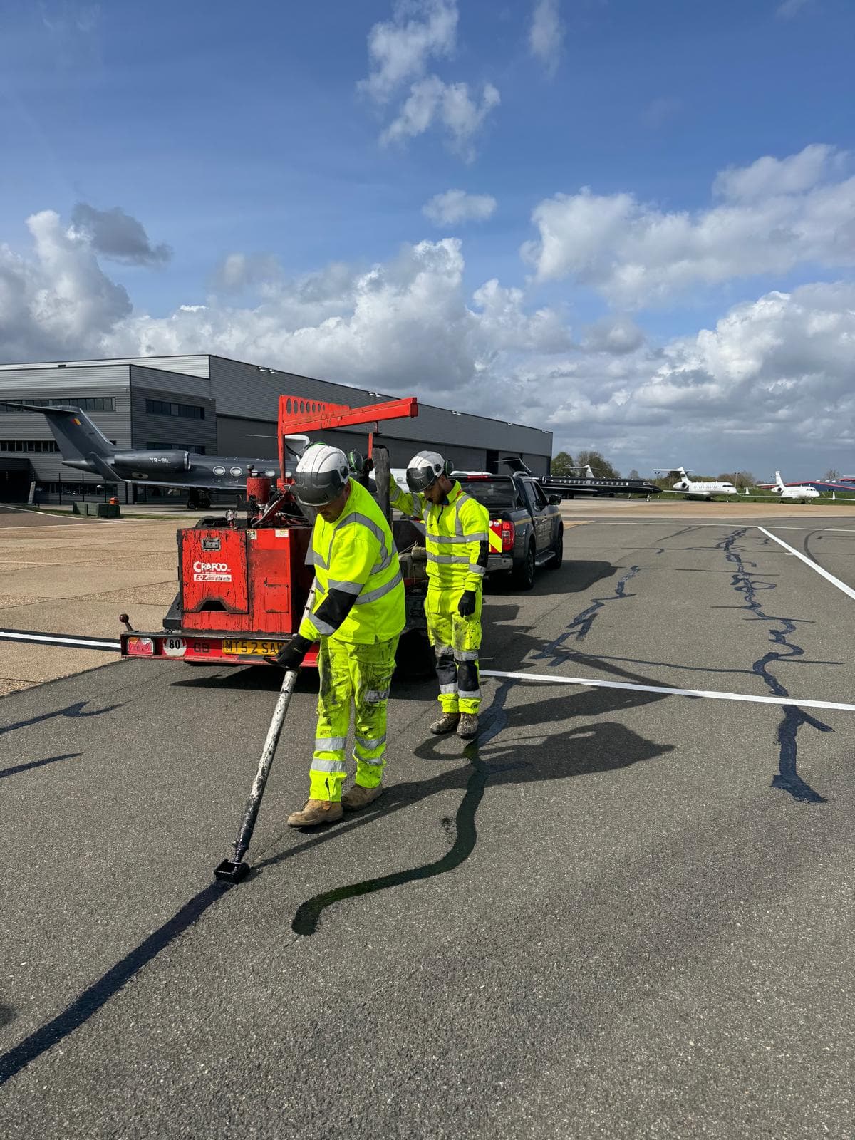 Two workers in hi-vis using a red crack-sealing machine on an airfield apron with parked jets and hangars behind
