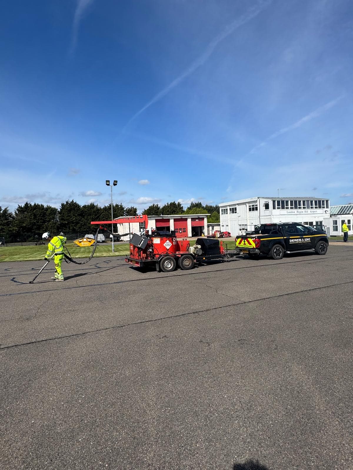 Worker in hi-vis using a lance on an airfield apron with a red trailer and branded Shepherd and Sons pickup at Biggin Hill