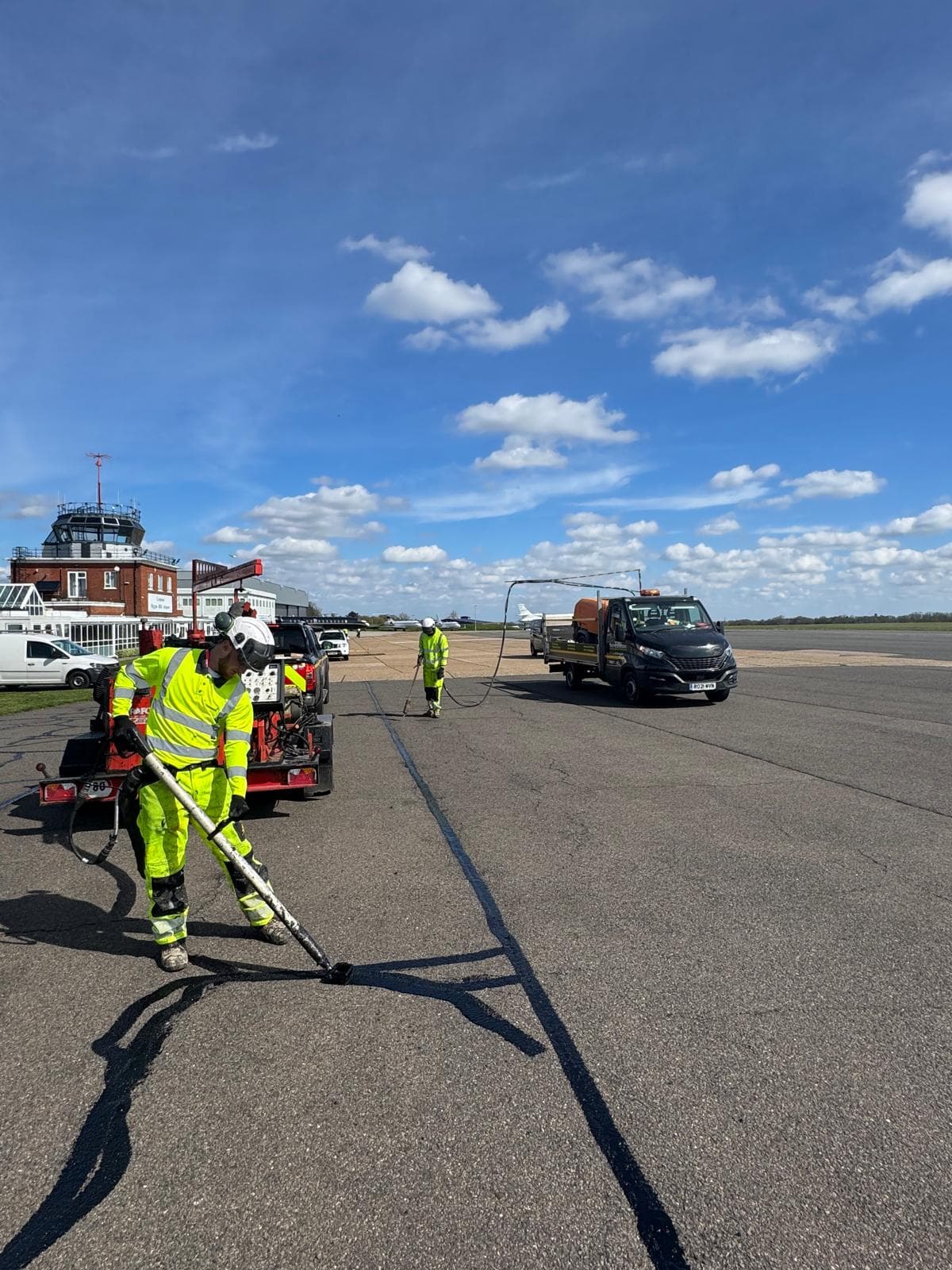 Workers in hi-vis carrying out crack-sealing on an airfield apron with work vans and a control tower behind