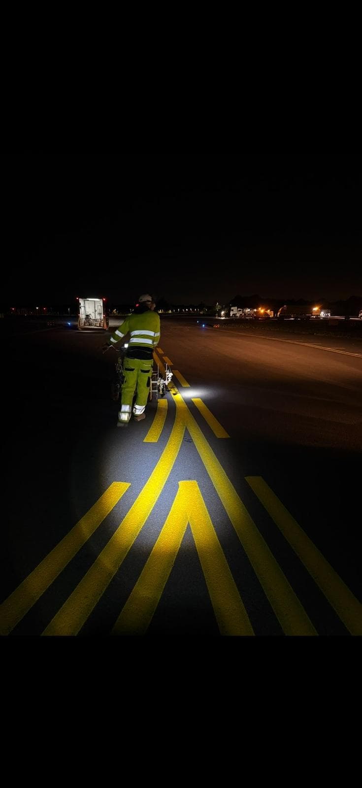 Operative in hi-vis pushing a line-marking machine along a runway at night, applying fresh reflective yellow markings