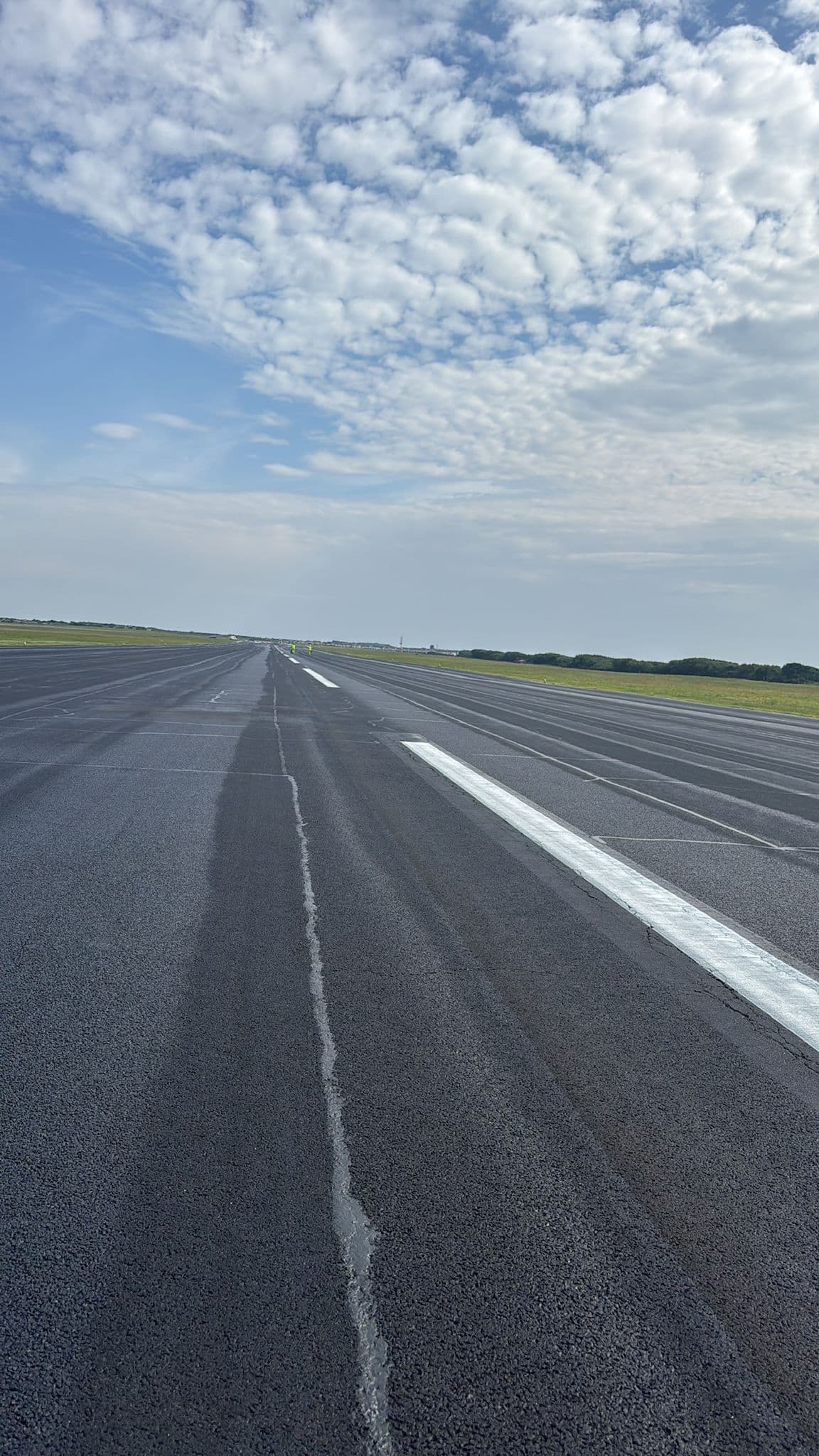 Wide view down a long runway under a big cloudy blue sky, with grass verges and workers in the distance