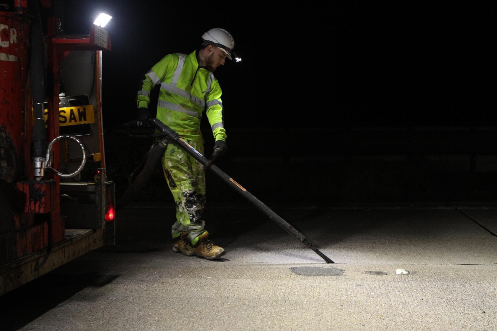 Worker in hi-vis using a long lance to seal a joint in a concrete road surface at night beside a work trailer