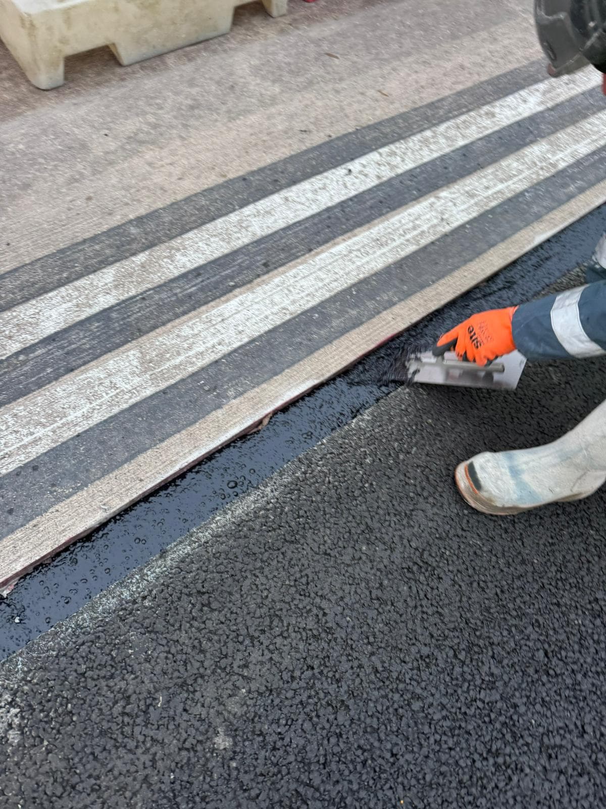 Gloved hands using a hand tool to lay black overbanding sealant along a road edge beside worn white markings