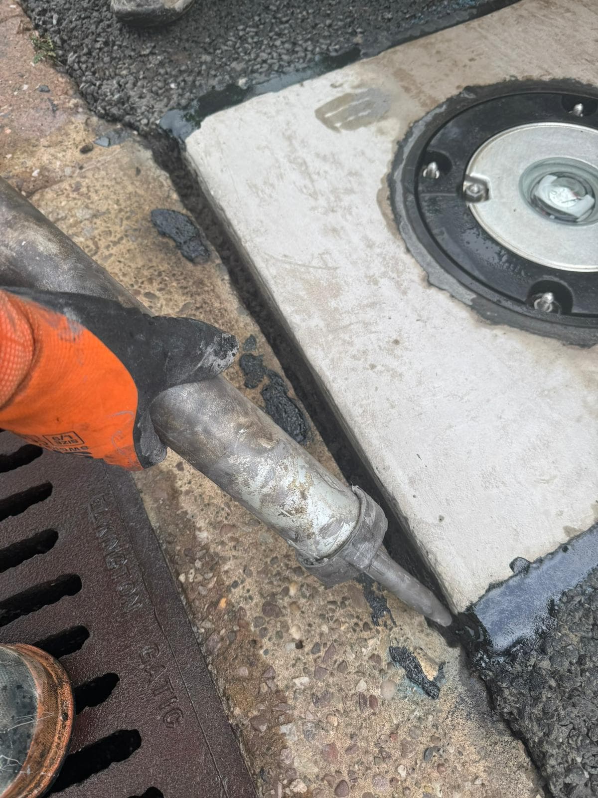 Gloved hand applying black sealant along the edge of a concrete slab beside a round metal access cover