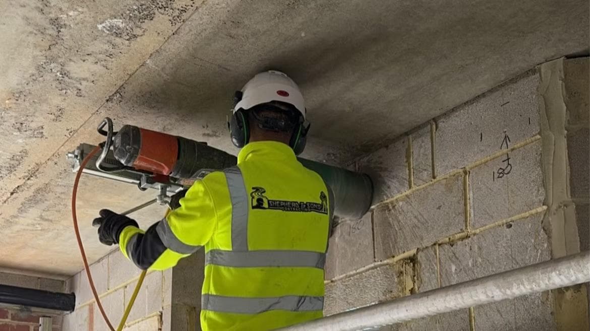 Worker in hi-vis and ear defenders operating a core-drilling rig to bore through a blockwork wall