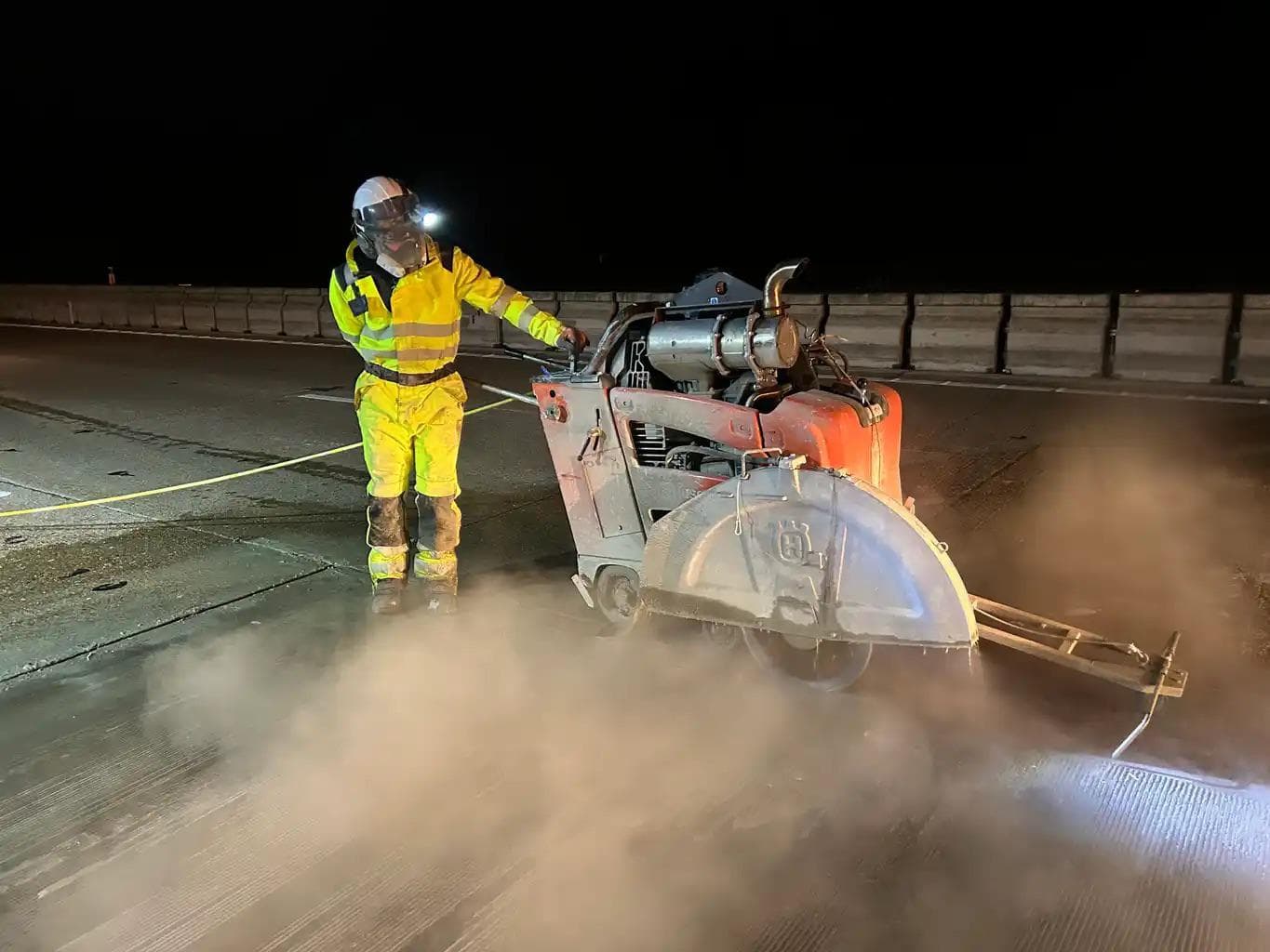 Worker in hi-vis operating a large walk-behind concrete floor saw at night with dust clouds around the blade