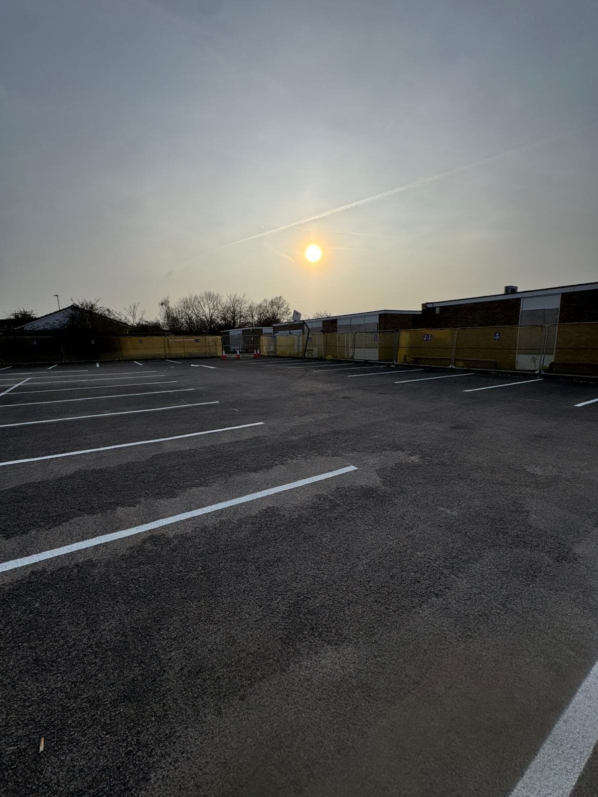 Empty freshly line-marked car park with white bay markings, temporary fencing and a low building at sunset