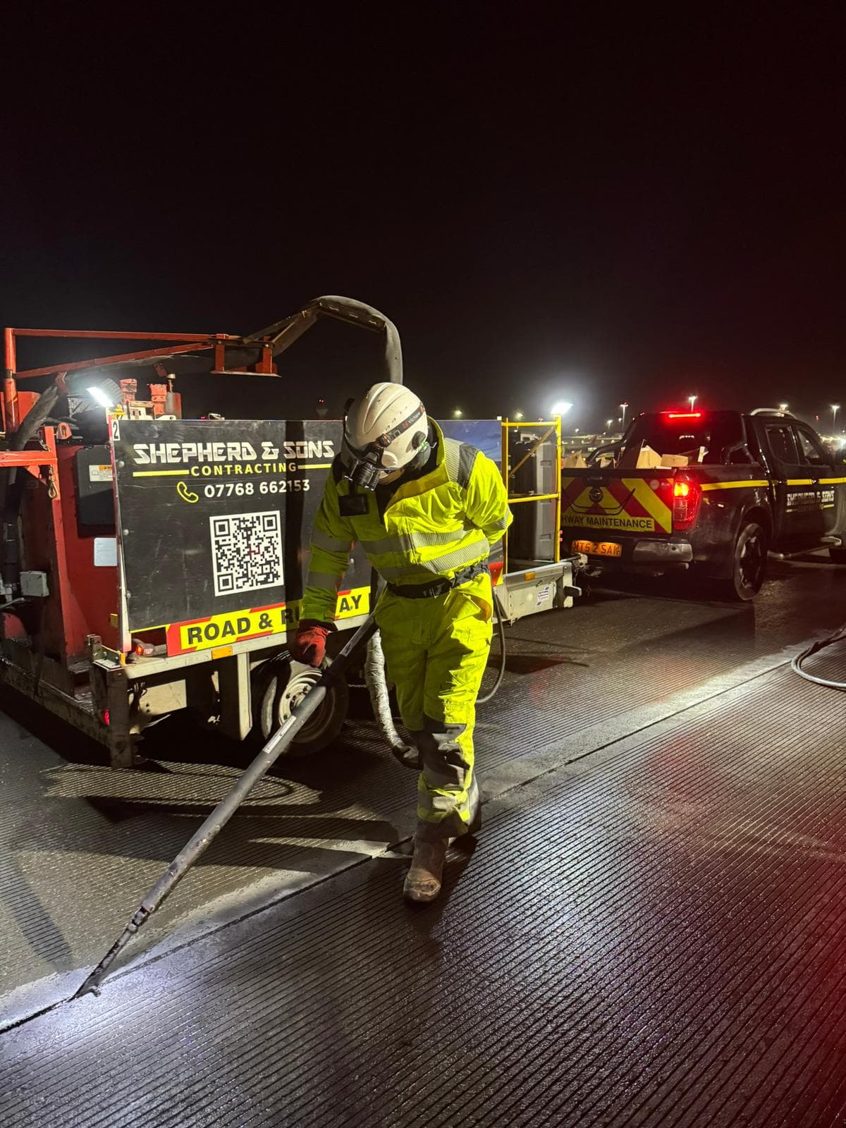 Operative in hi-vis and respirator using a handheld cut-off saw at night beside a Shepherd and Sons Contracting trailer