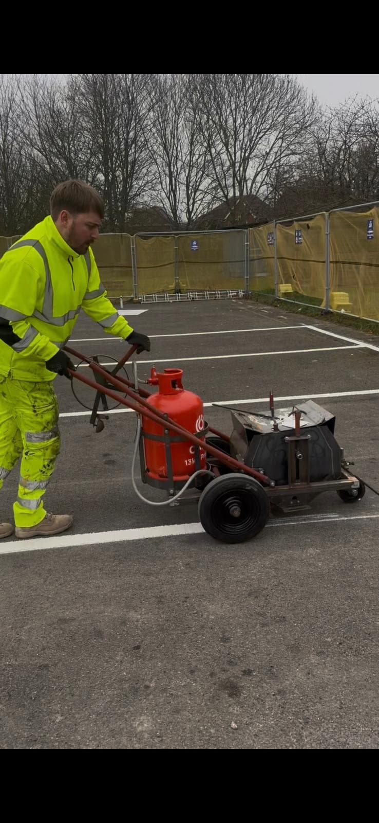 Worker in hi-vis pushing a gas-powered line-marking machine across a car park with fresh white bay lines
