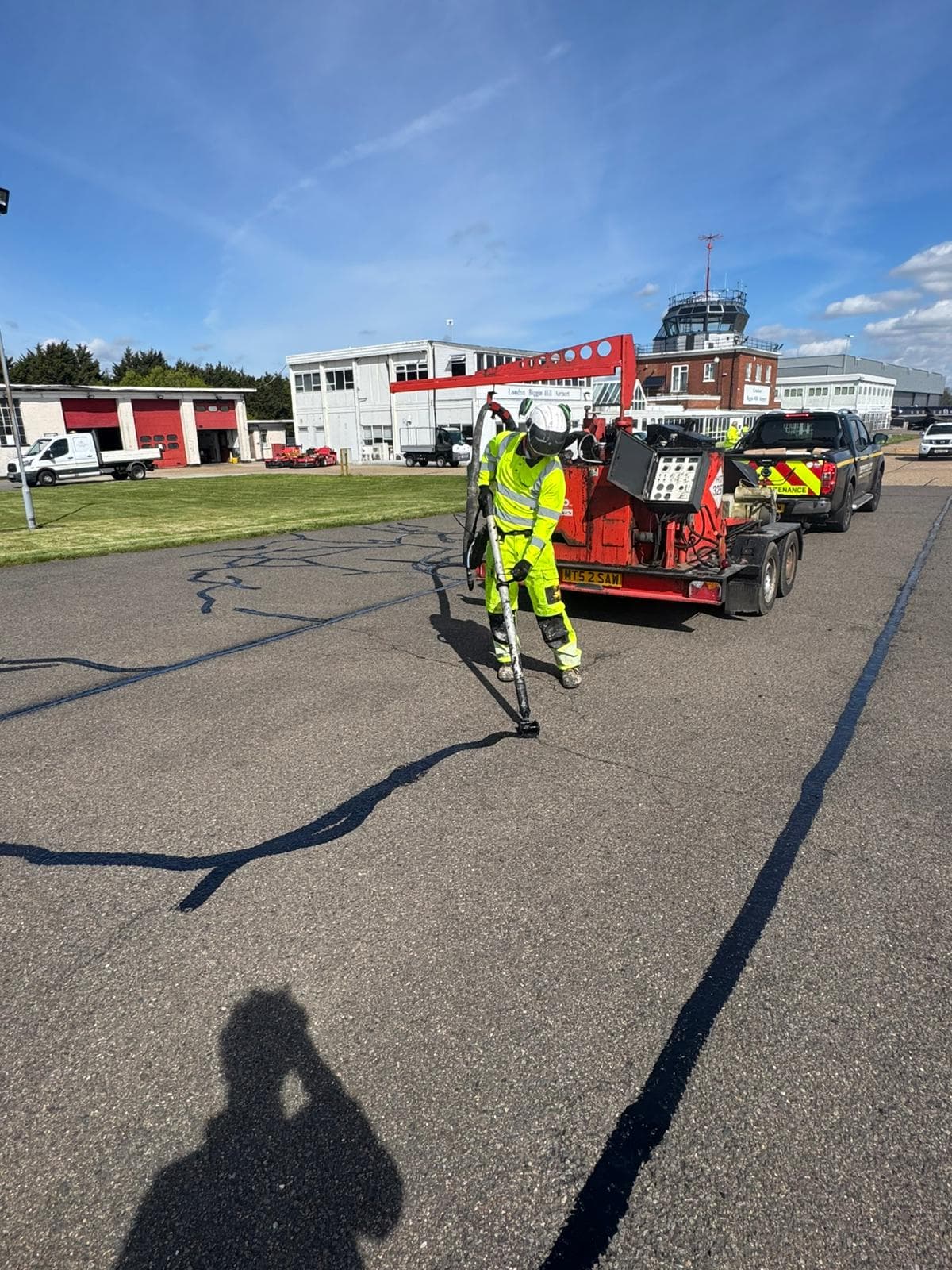 Worker in hi-vis using a hand lance to seal cracks across a tarmac area with a red crack-sealing trailer behind