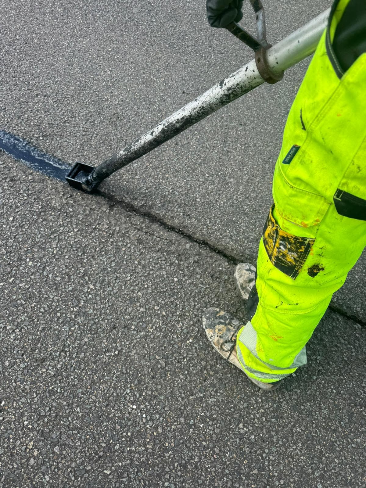 Close-up of an operative using a hand lance to draw black crack sealant along a crack in grey asphalt