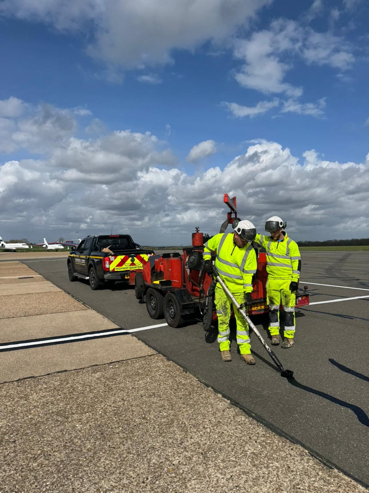 Two workers in hi-vis operating a red crack-sealing trailer on a tarmac apron with a pickup and aircraft behind