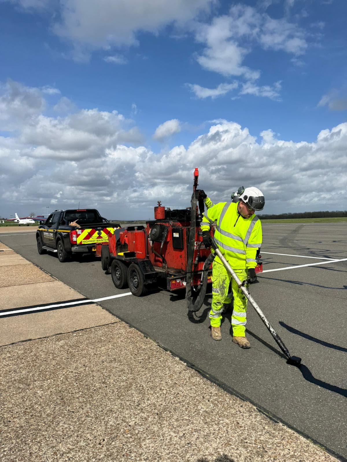 Worker in hi-vis operating a red trailer-mounted crack-sealing machine on a tarmac apron with a pickup behind