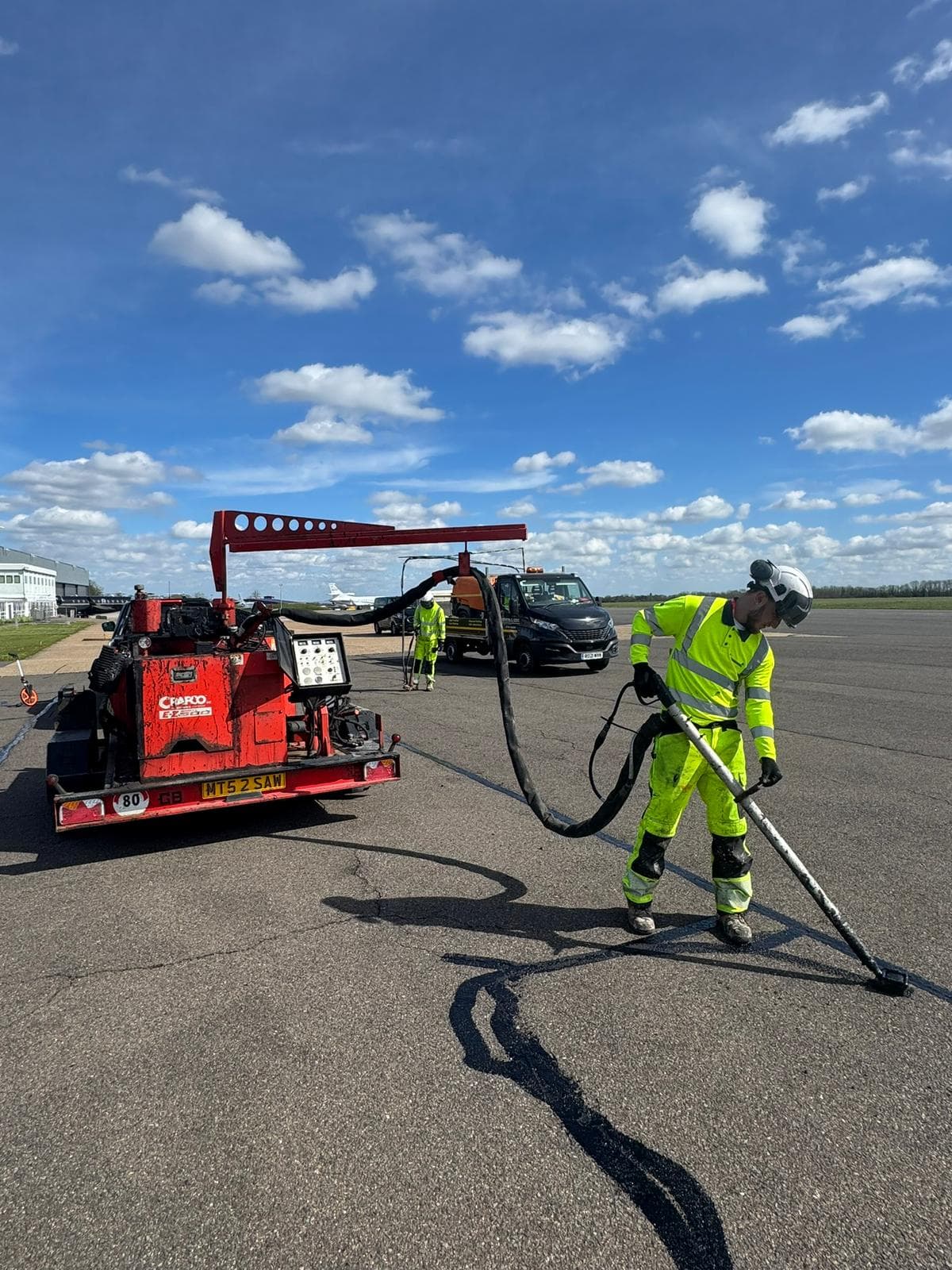 Worker in hi-vis using a hand lance from a red crack-sealing machine on a wide tarmac area under a blue sky