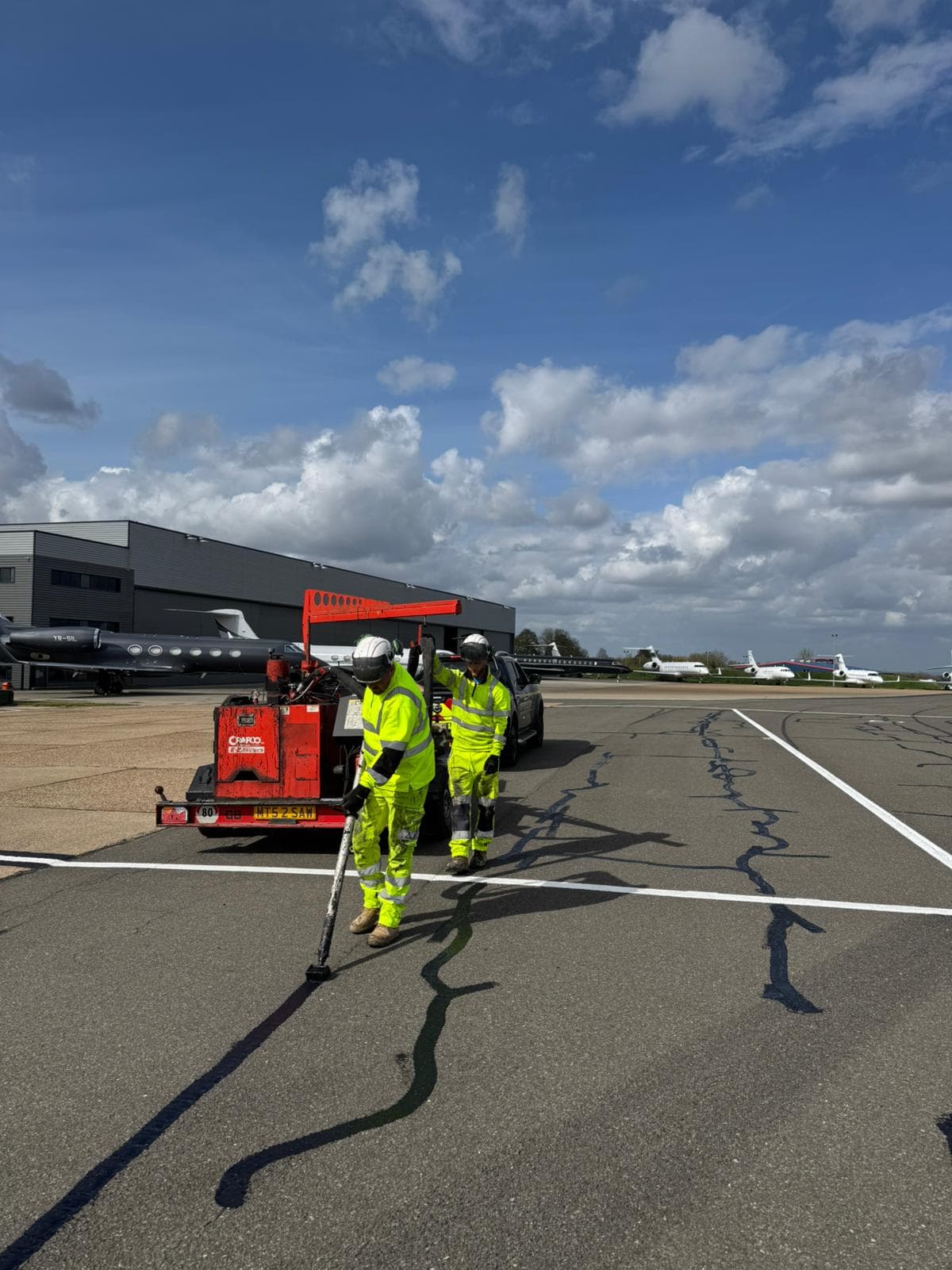 Two workers in hi-vis operating a red crack-sealing machine on a tarmac apron with parked aircraft and a hangar behind