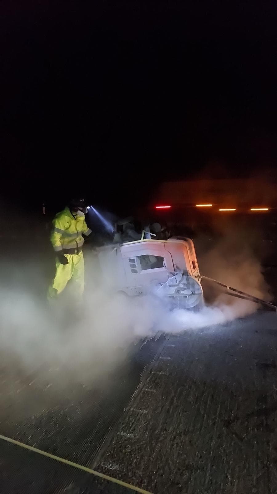 Operative in hi-vis with a head torch operating a floor saw at night amid dust with warning lights behind
