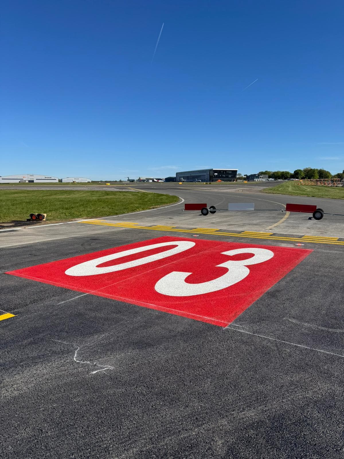 Large white 03 runway designation number painted on a red surface at a runway threshold with hangars behind