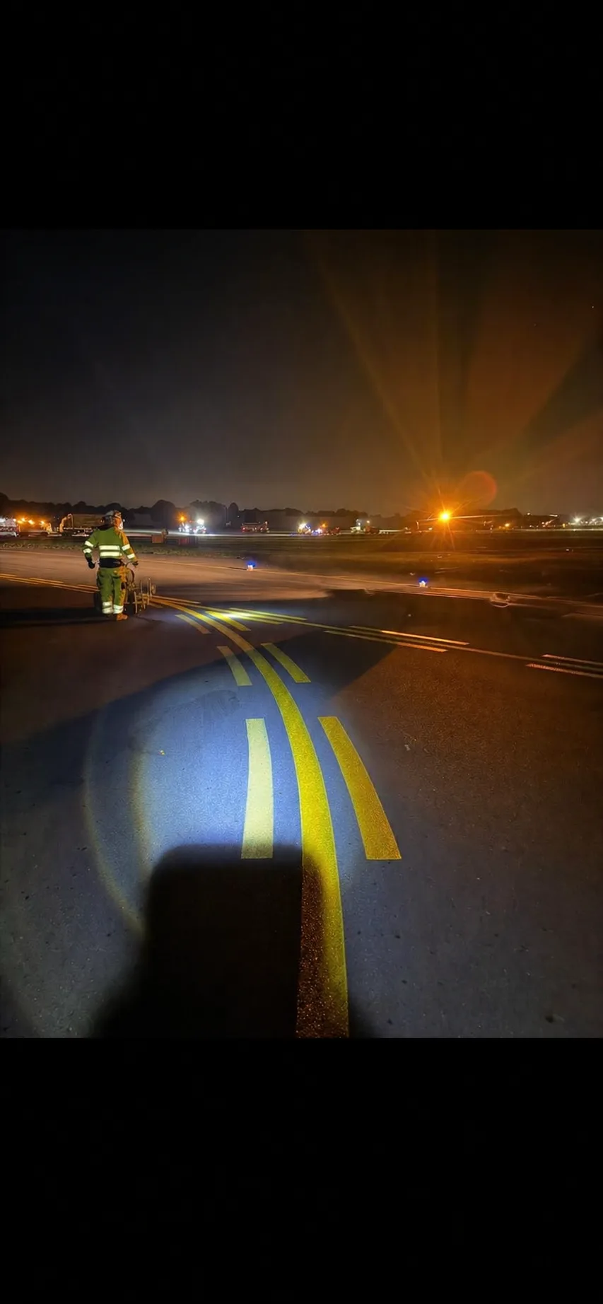 Freshly applied yellow taxiway lines picked out by a work light at night, with an operative and marking trolley beyond