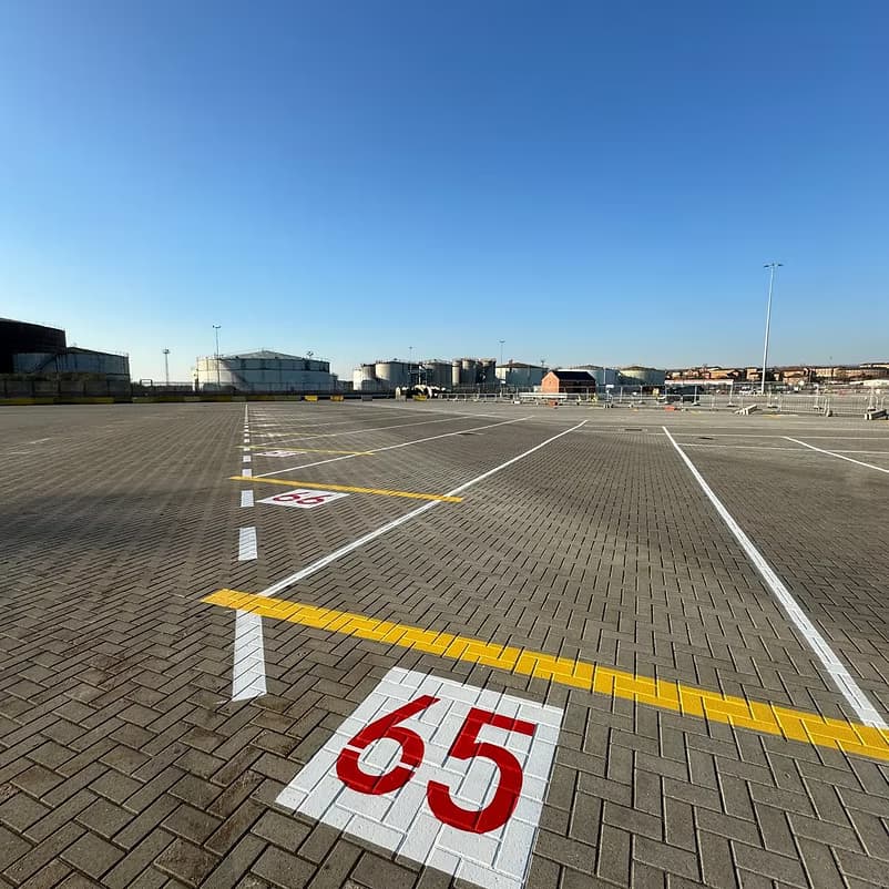 Block-paved apron with a large red 65 bay number and white and yellow line markings, storage tanks in the distance