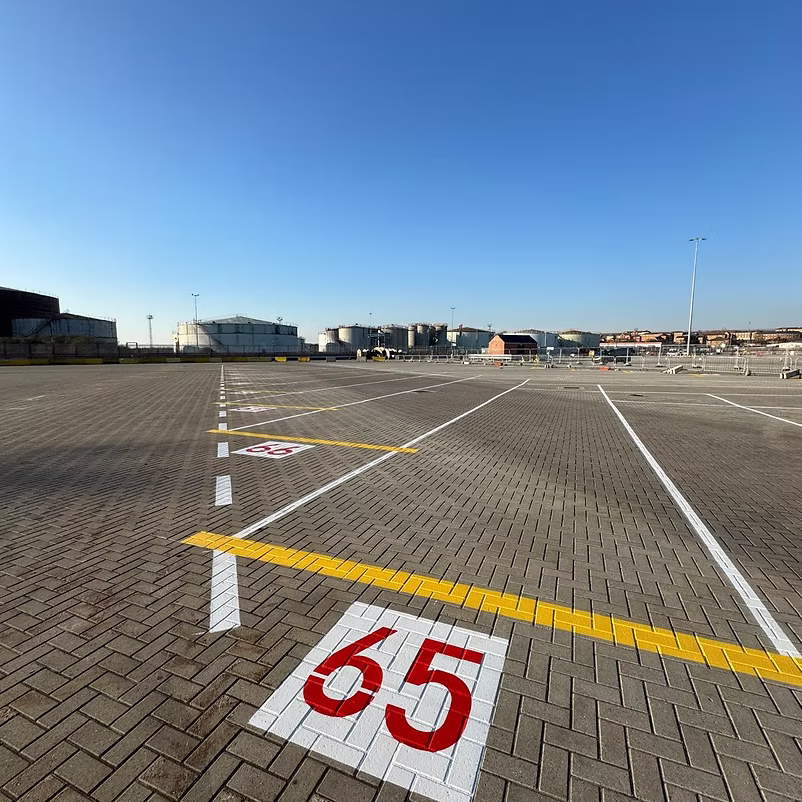 Block-paved apron with a large red 65 bay number and white and yellow line markings, storage tanks in the distance