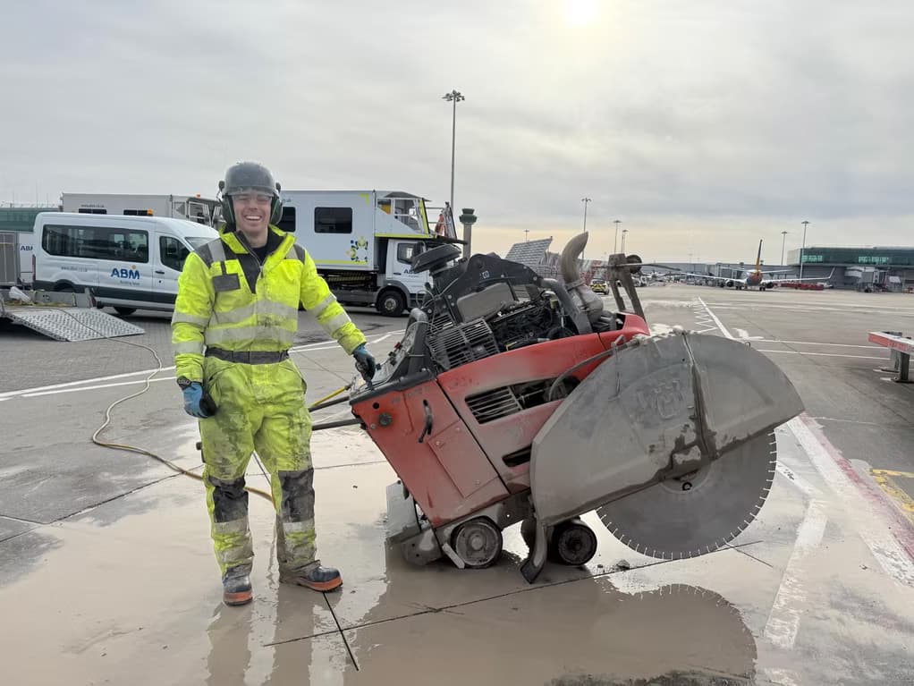 Worker in yellow hi-vis and helmet beside a large red road saw on an airport apron with aircraft behind