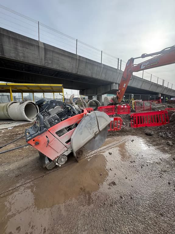 Red road saw on a muddy construction site beneath a bridge with an excavator, concrete pipes and barriers