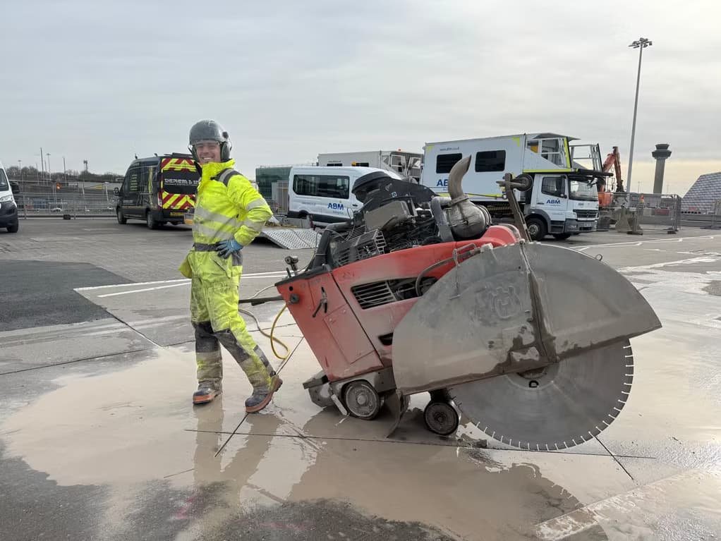 Worker in yellow hi-vis and helmet beside a large red road saw on an airport apron with vehicles behind