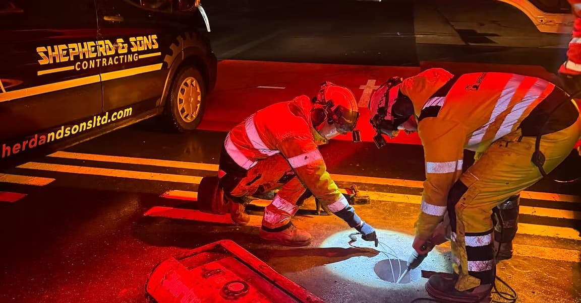 Two workers in hi-vis kneeling at night over an open inspection chamber in red light beside a company van