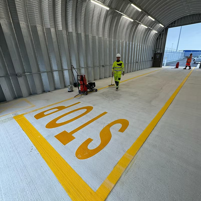 Worker in green hi-vis sweeping a large yellow STOP marking on a concrete surface in a covered walkway