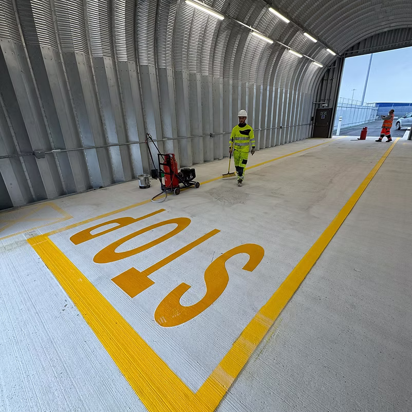 Worker in green hi-vis sweeping a large yellow STOP marking on a concrete surface in a covered walkway