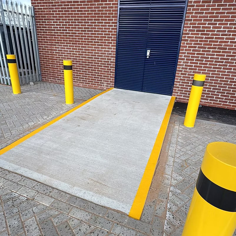 Concrete ramp with fresh yellow edge lines and safety bollards in front of a blue shutter door on a brick building