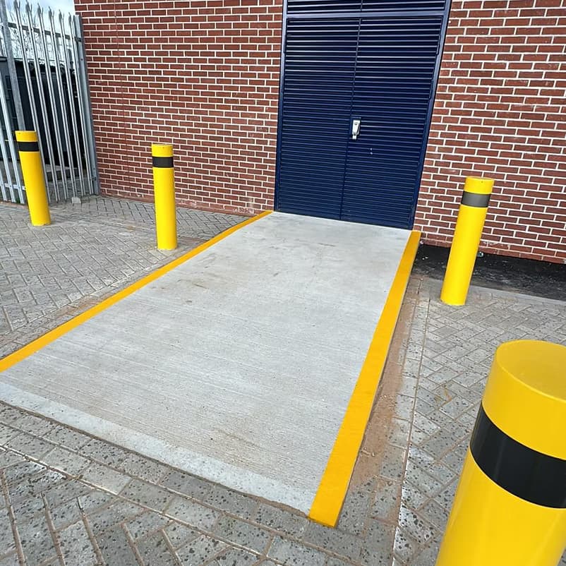 Concrete ramp with fresh yellow edge lines and safety bollards in front of a blue shutter door on a brick building