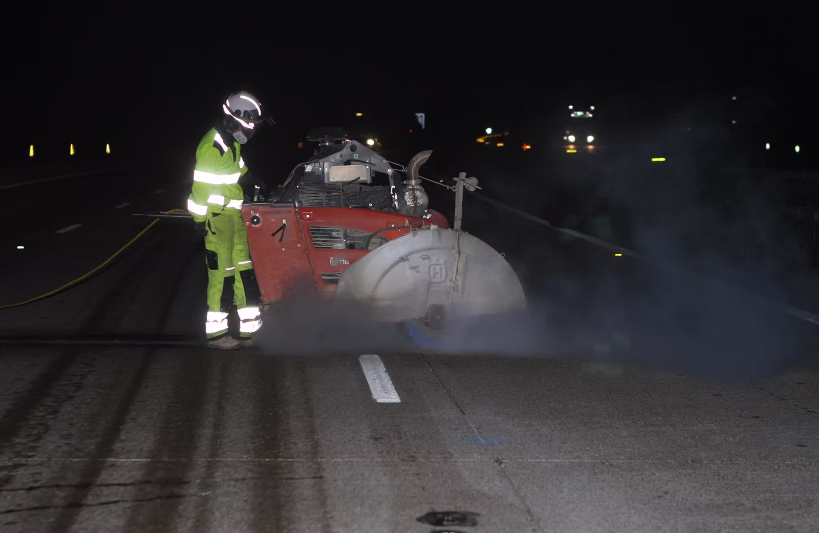 Worker in green hi-vis and helmet operating a red floor saw on a road at night with dust around the blade