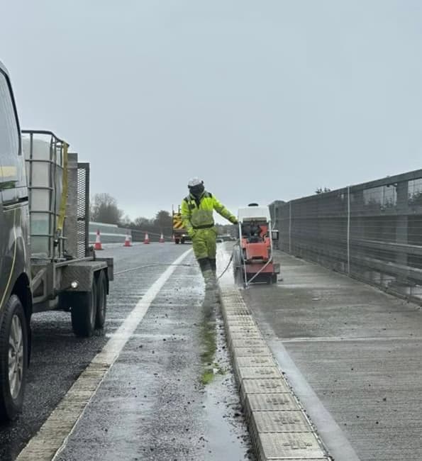 Worker in yellow hi-vis using a lance beside a small machine on a wet road with a slot drain and crash barrier