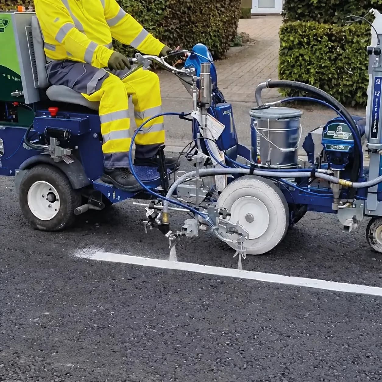 Operator in hi-vis riding a blue road line-marking machine applying a white line onto a dark asphalt surface