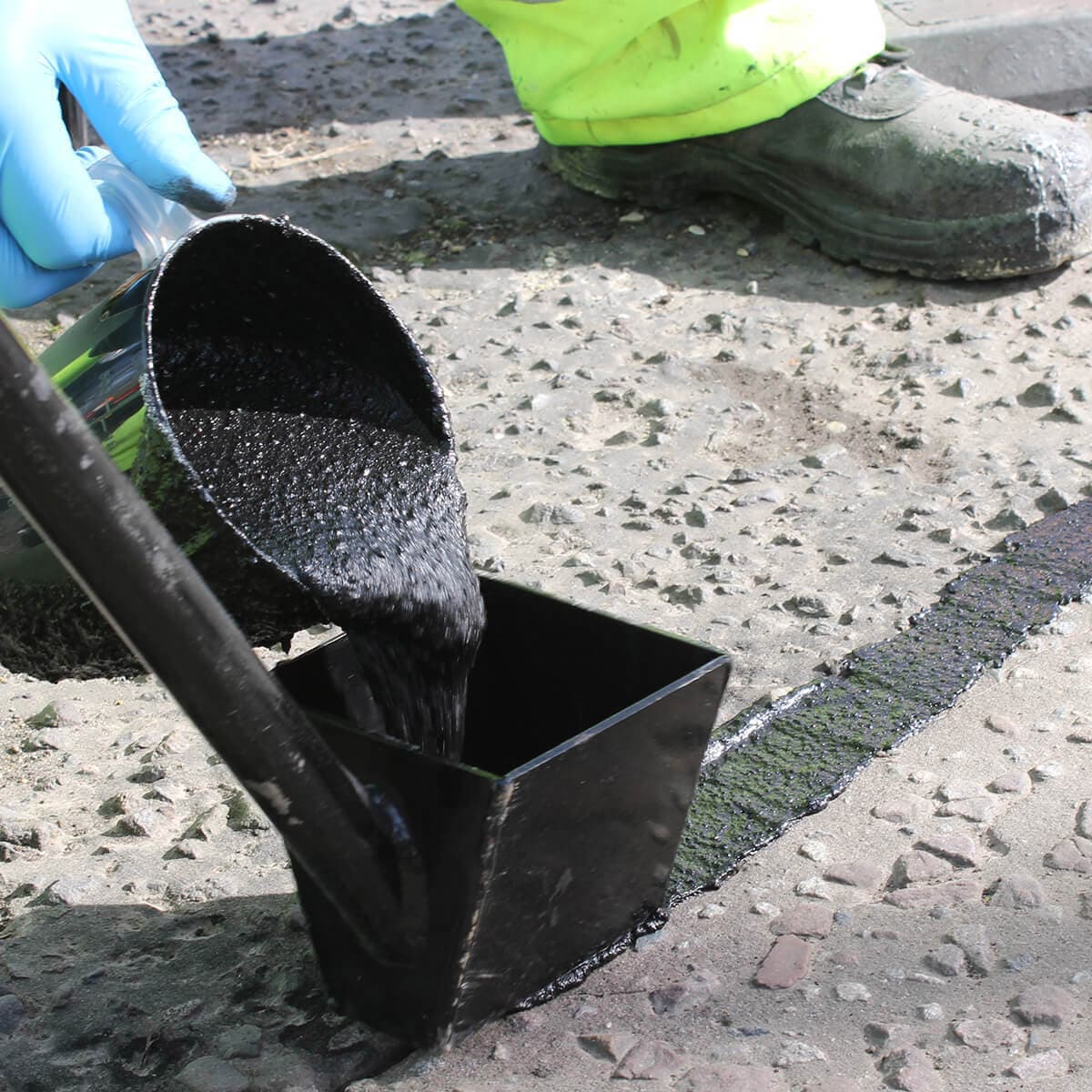 Black bituminous overbanding compound being poured from a bucket into an applicator box on a road surface