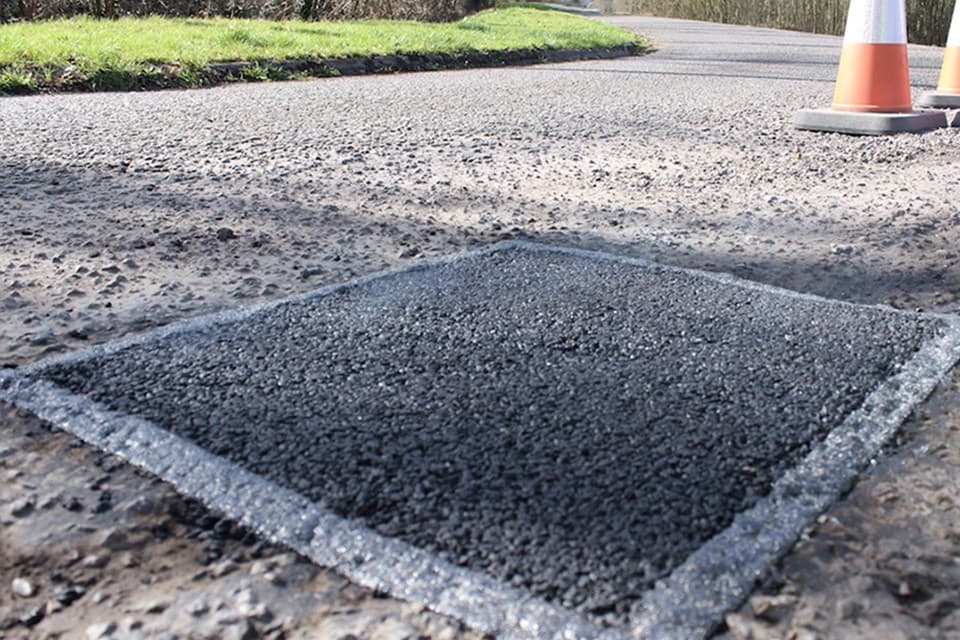 Square patch of fresh dark asphalt laid into a repaired section of pavement surrounded by lighter aggregate
