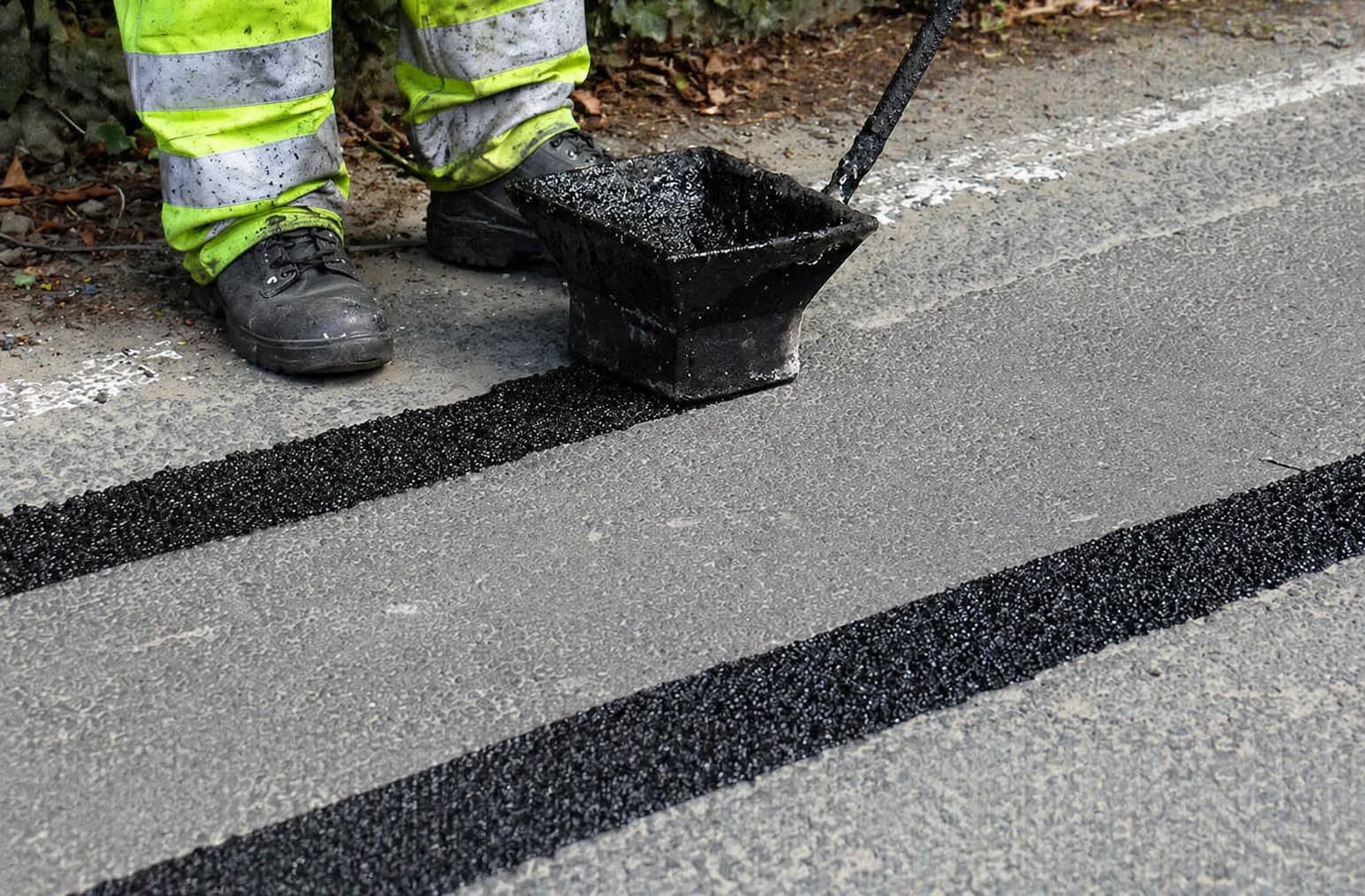 Black overbanding strips being laid across a grey road surface using an applicator box beside a worker in hi-vis