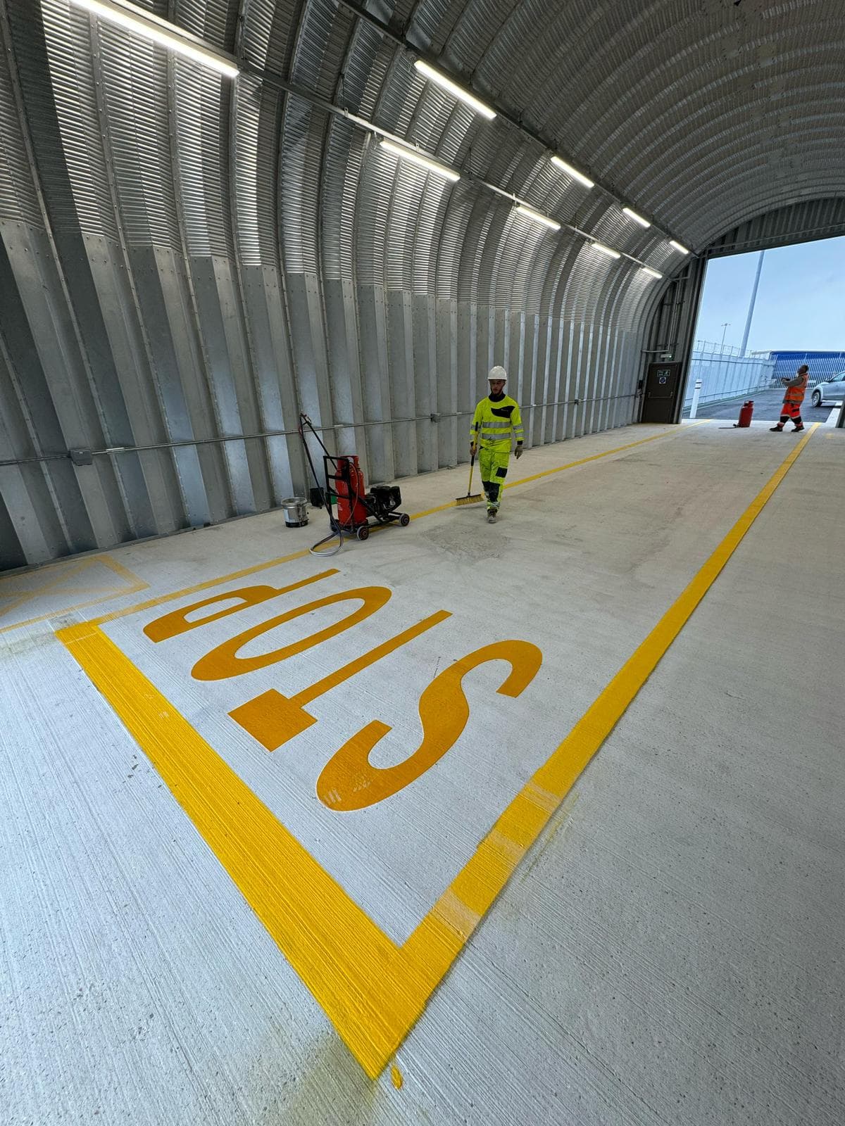 Worker in hi-vis with a broom on a concrete floor with a yellow STOP marking inside a curved metal-clad tunnel