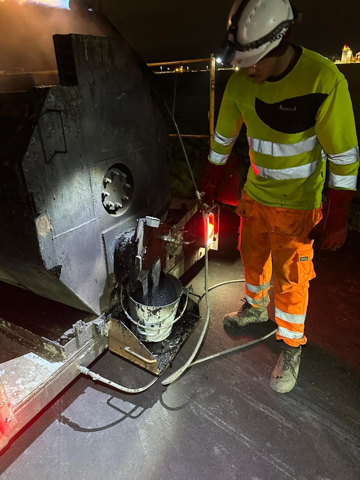 Worker in hi-vis and hard hat drawing hot sealant from a black melter unit at night