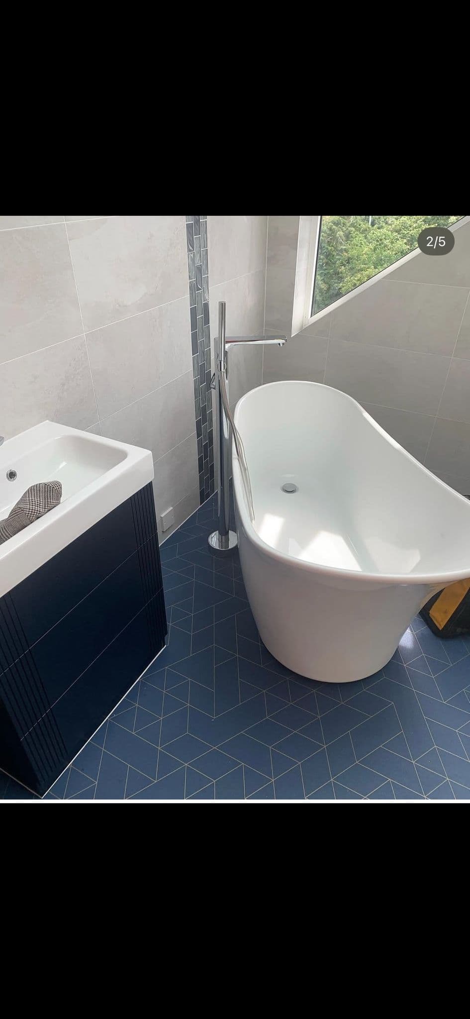 Modern bathroom with a freestanding white slipper bath, grey tiled walls, dark herringbone floor and a dark vanity unit