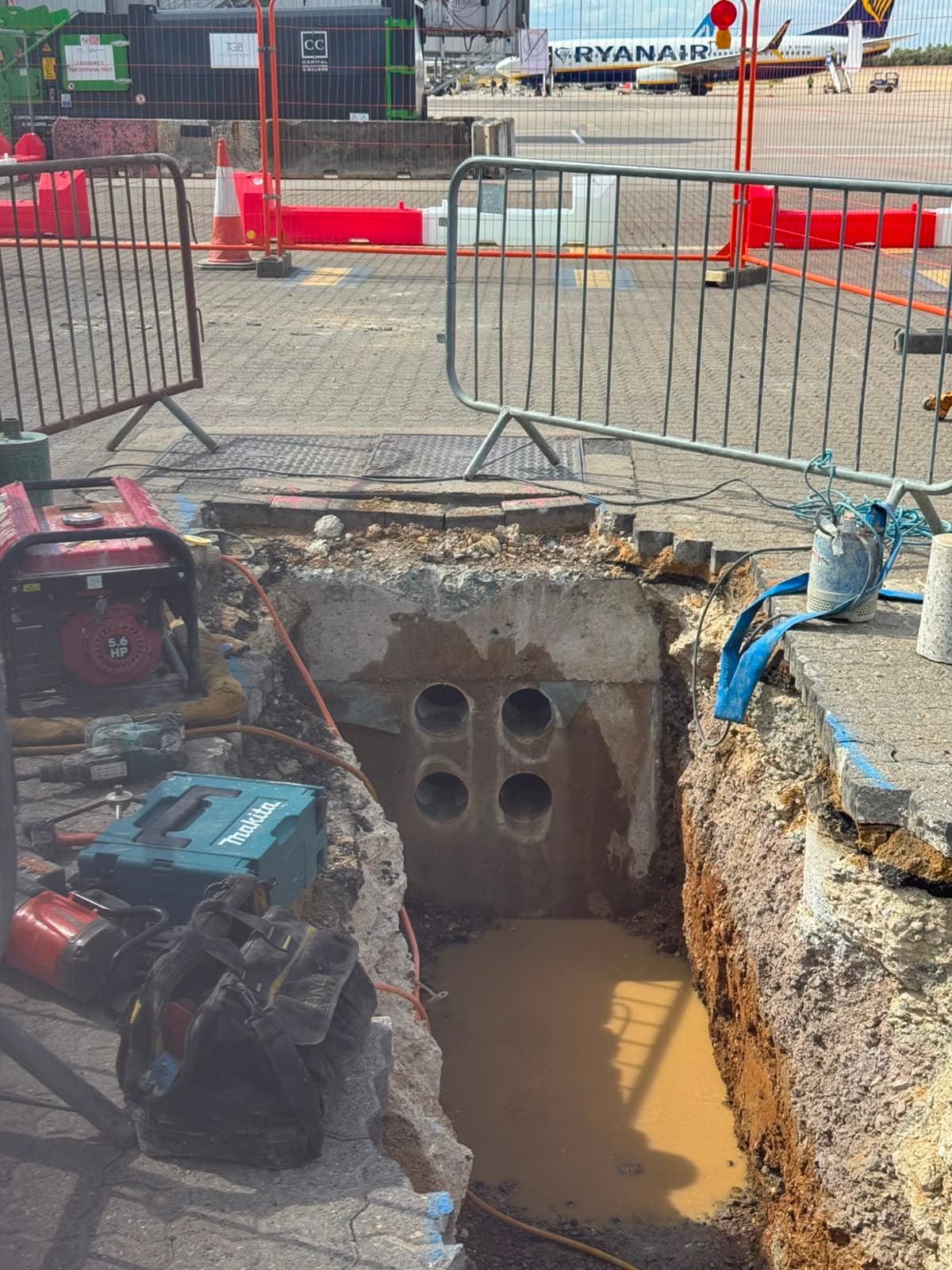 Four cored holes through a concrete duct block in an open excavation, with an aircraft on the apron behind