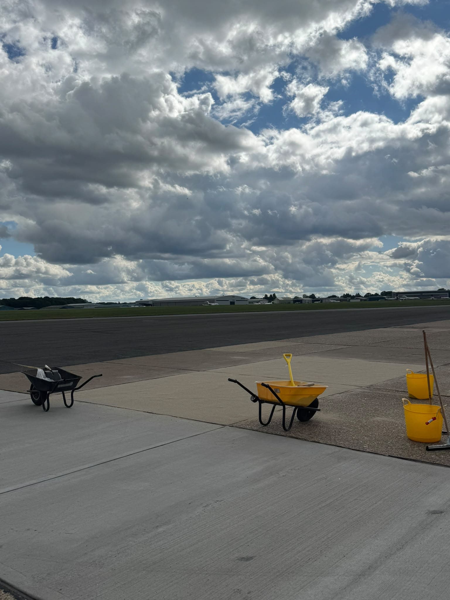 Barrows, buckets and hand tools staged neatly together on an airfield apron beside the working area, with the runway and hangars behind