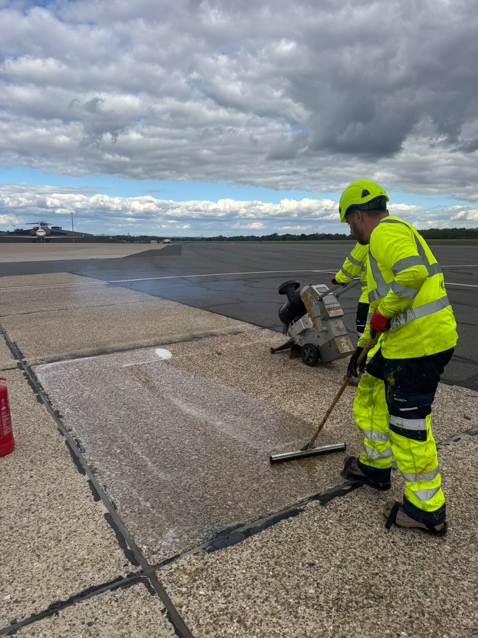 An operative spreading resin across a prepared concrete bay with a squeegee on an airfield apron, the surface planer standing by and the runway beyond